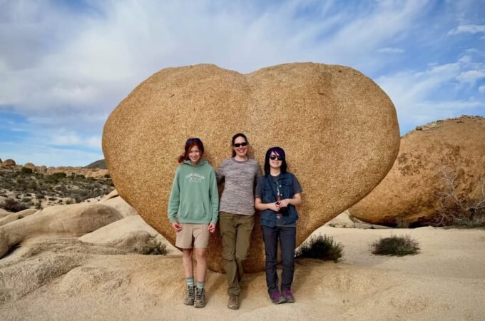 A group of three travelers posing in front of a massive heart-shaped boulder in a desert landscape.