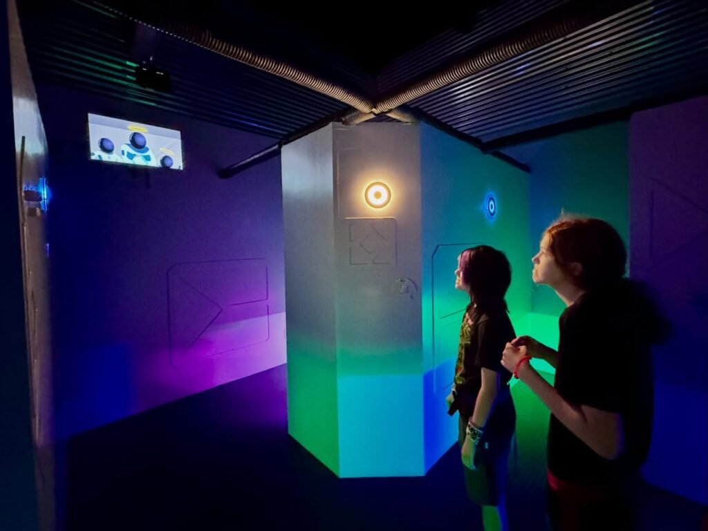 A collage showing a colorful laser tag arena with illuminated walls. Two people are playing the game while a screen monitors their scores.