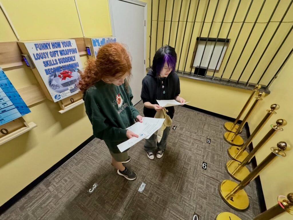 Two people with papers stand in a room with barred windows and informational posters. Gold stanchions and wall displays are visible.