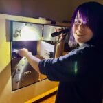 Heart Rock A person with purple hair uses a flashlight to search a wall panel during an escape room experience.
