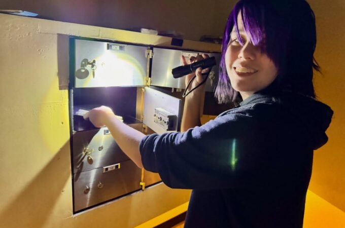 A person with purple hair uses a flashlight to search a wall panel during an escape room experience.