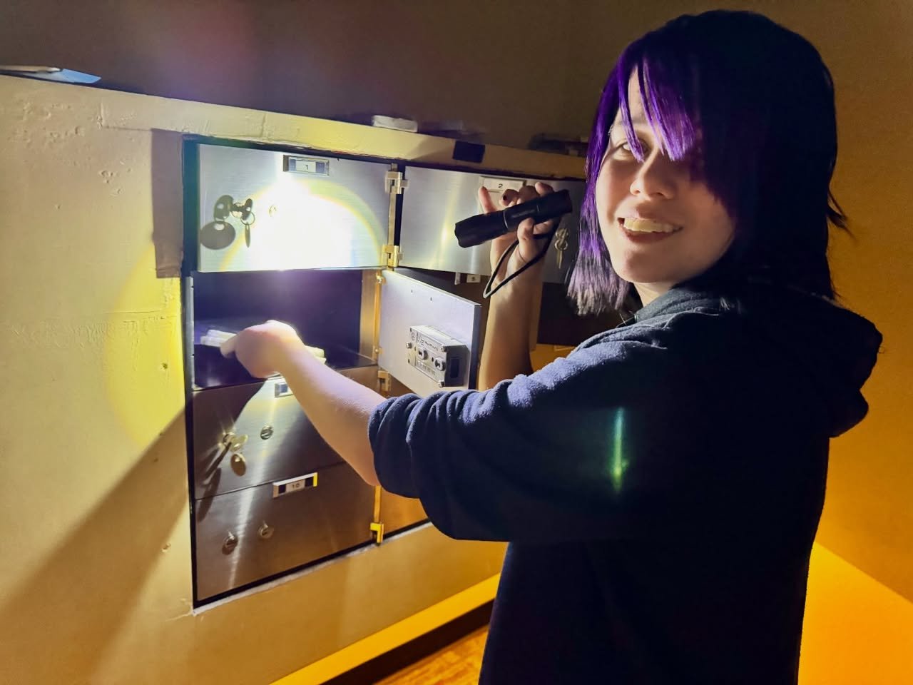A person with purple hair uses a flashlight to search a wall panel during an escape room experience.