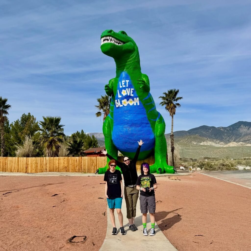 Three people stand in front of a large inflatable green dinosaur with the phrase 'Let Love Bloom' in a desert landscape under a clear sky.