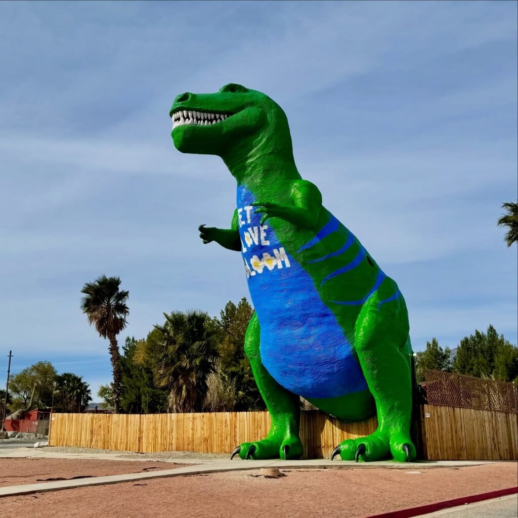 A large green T-Rex statue standing in an outdoor area with palm trees in the background.