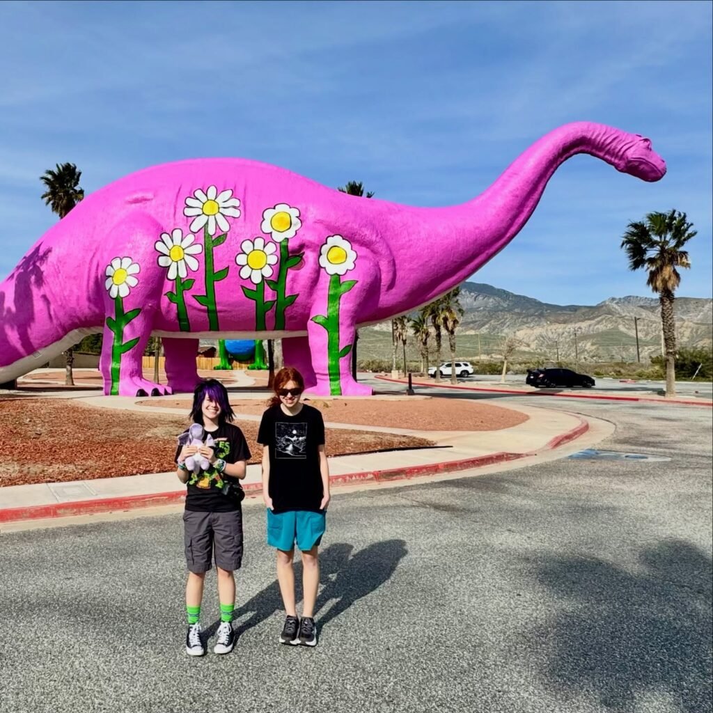 Two visitors pose in front of a large, vibrant pink dinosaur sculpture decorated with daisies under a clear blue sky.