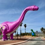 A large pink dinosaur sculpture stands next to a smaller green one in a park with palm trees.
