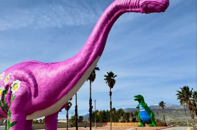 A large pink dinosaur sculpture stands next to a smaller green one in a park with palm trees.