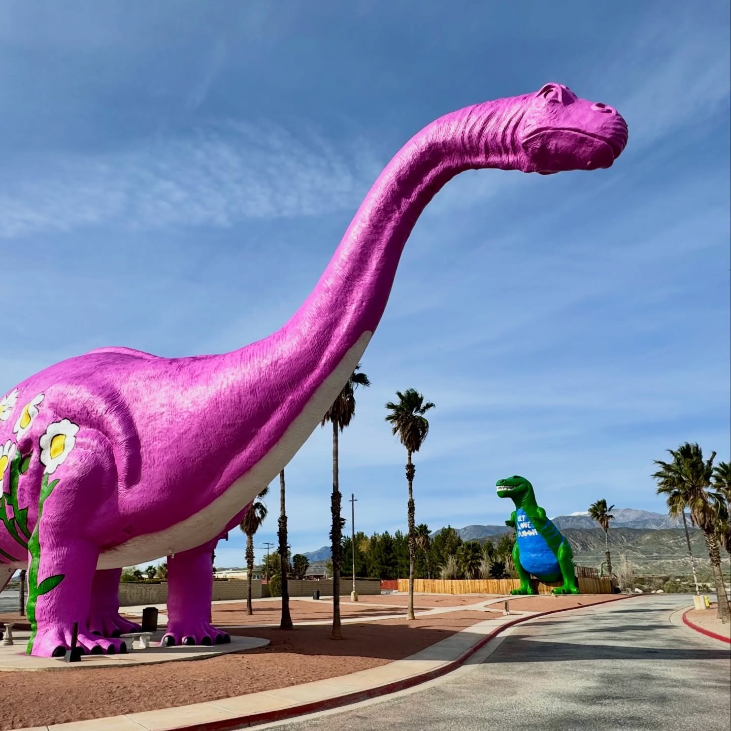 A large pink dinosaur sculpture stands next to a smaller green one in a park with palm trees.
