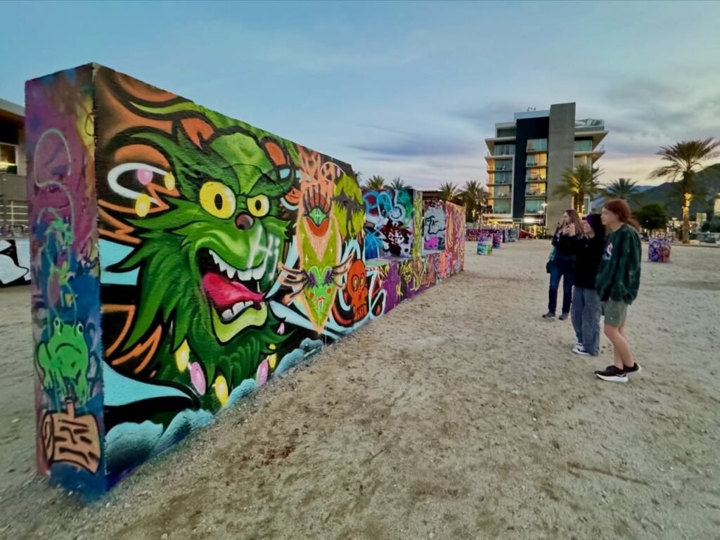 A large colorful mural featuring a fierce green face decorates a long urban wall in a parking area.