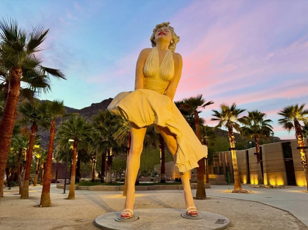 A large golden statue of Marilyn Monroe in her iconic dress, standing amidst palm trees as the sky turns pink and purple at dusk.