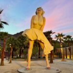 Emerald City Comic Con A large golden statue of Marilyn Monroe in her iconic dress, standing amidst palm trees as the sky turns pink and purple at dusk.