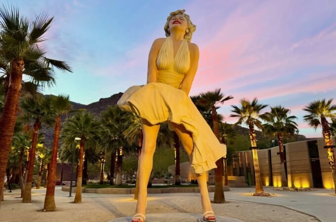 Downtown Palm Springs A large golden statue of Marilyn Monroe in her iconic dress, standing amidst palm trees as the sky turns pink and purple at dusk.