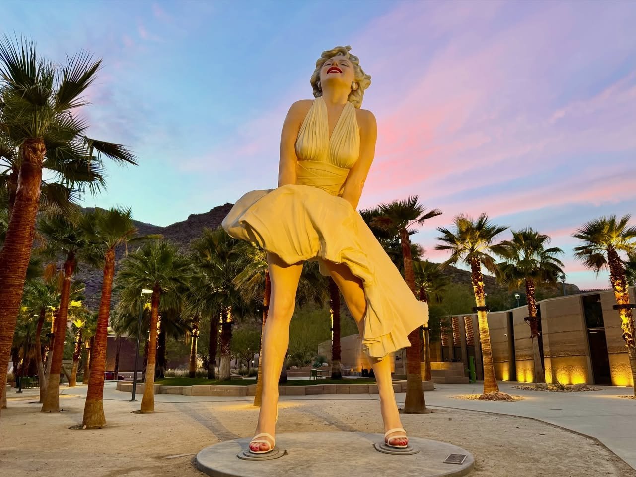 A large golden statue of Marilyn Monroe in her iconic dress, standing amidst palm trees as the sky turns pink and purple at dusk.