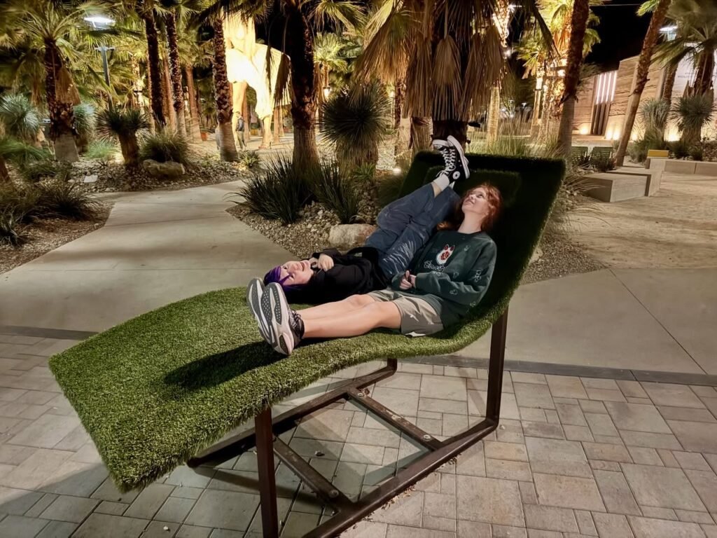 A person relaxes on a green lounge chair outdoors at dusk, surrounded by palm trees and a paved walkway.