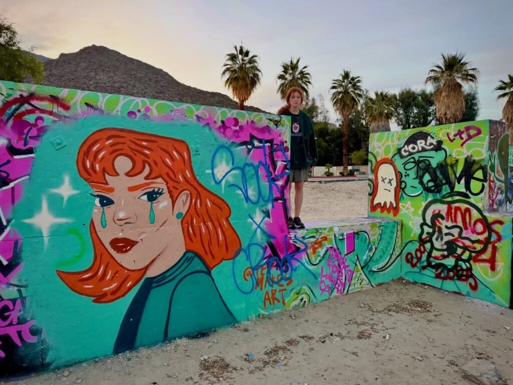 A person stands near low walls covered in bright teal, magenta, and turquoise graffiti beneath palm trees and mountains.