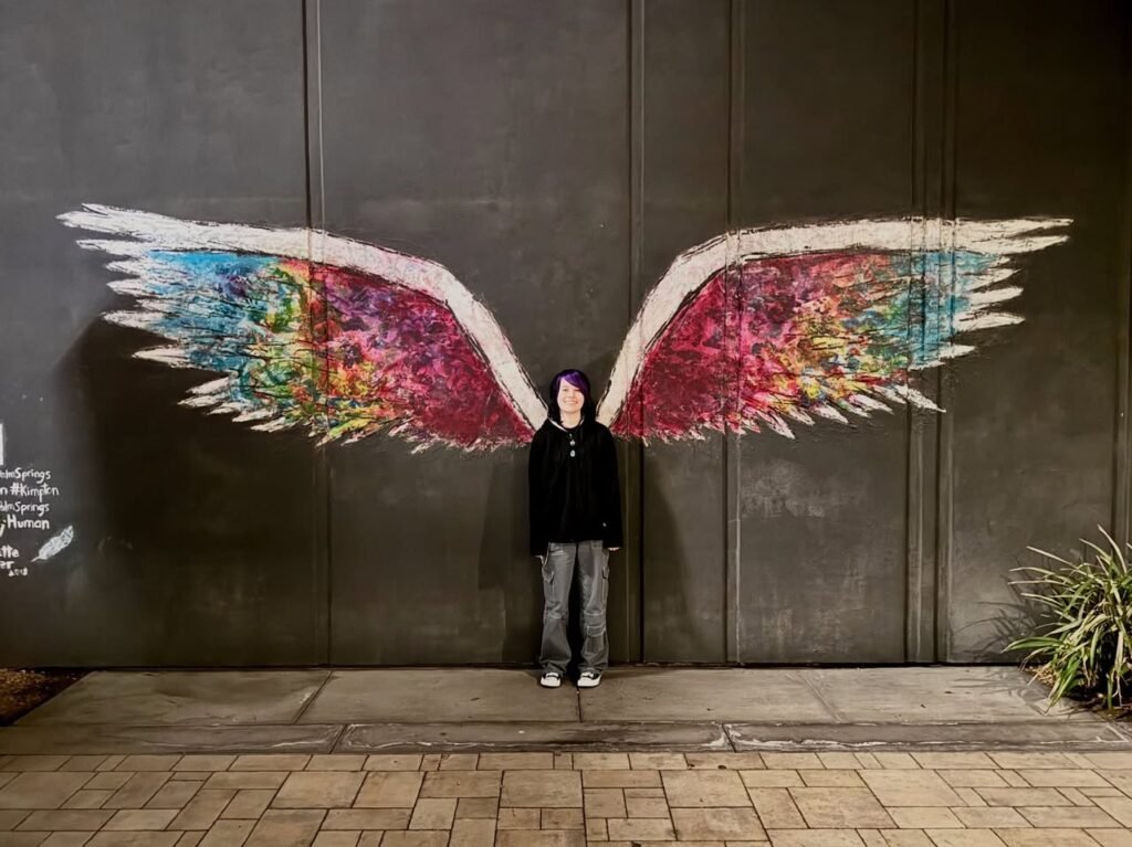 A person stands in front of a large, colorful winged angel mural on a wooden wall. The artwork features intricate, multicolored feathers that create a striking visual effect.