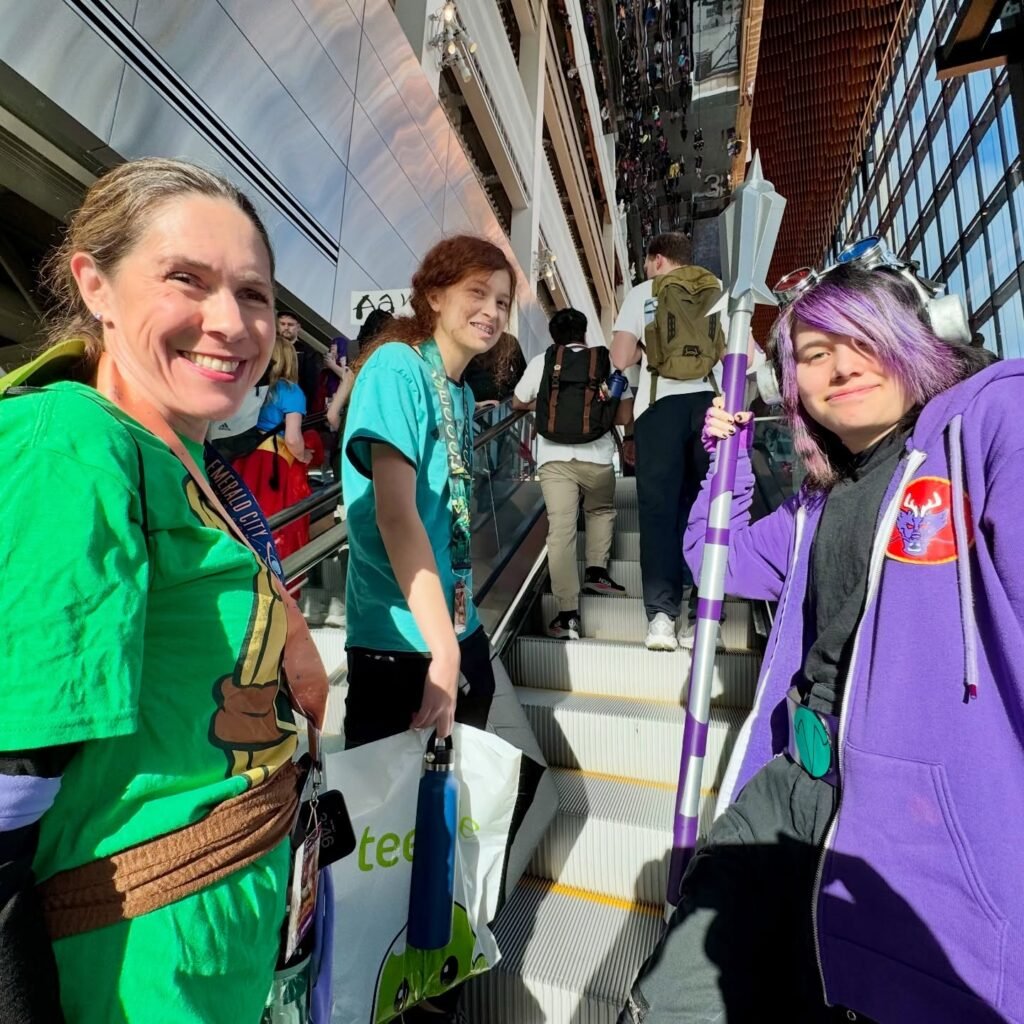 Three individuals pose together at a convention, wearing vibrant apparel and carrying themed props inside a large exhibition hall.