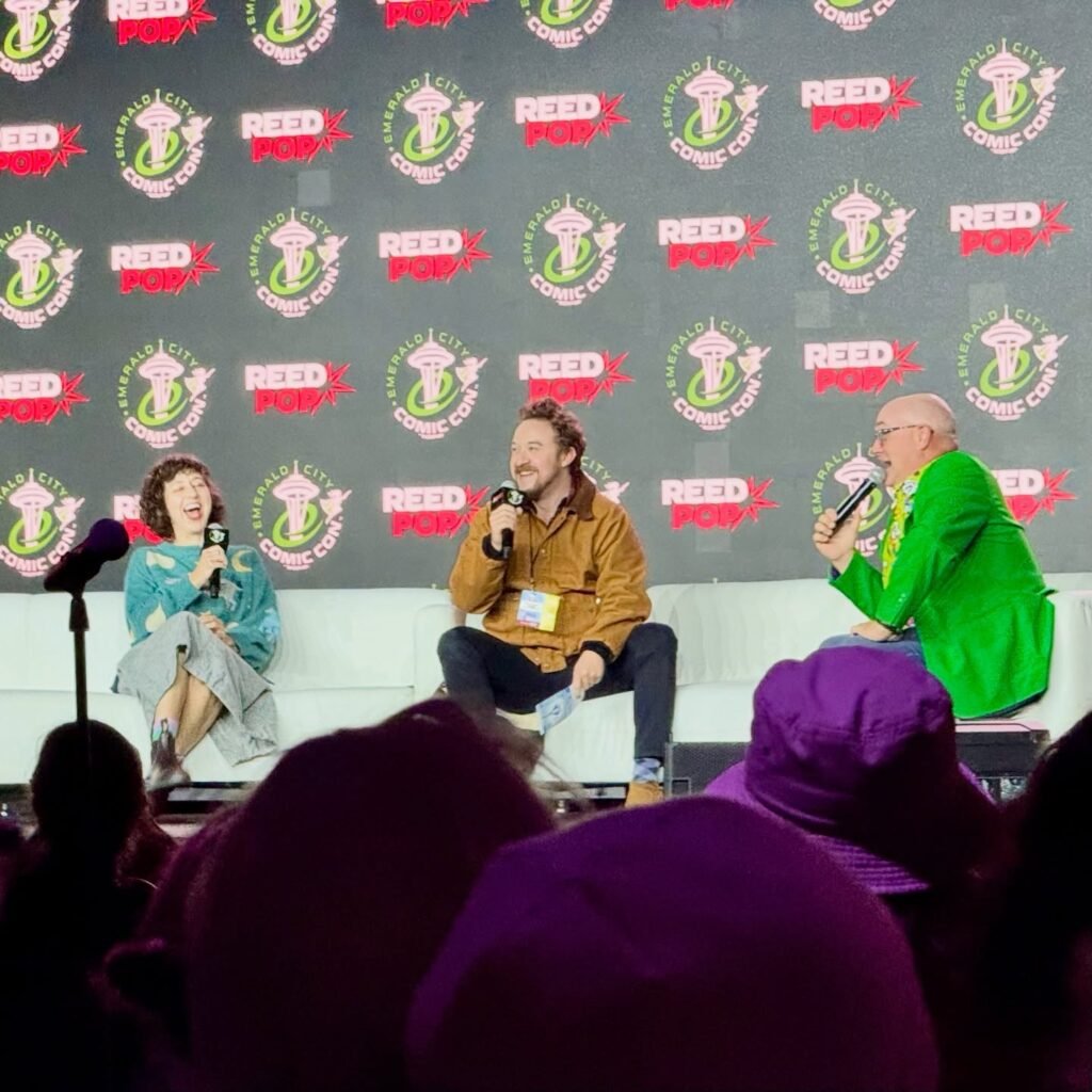 Panelists seated on stage at a comic convention with a branded backdrop