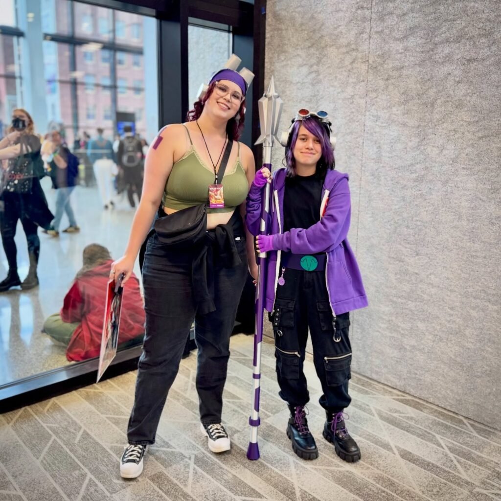 Two individuals in purple and green cosplay costumes hold props in a convention hall.