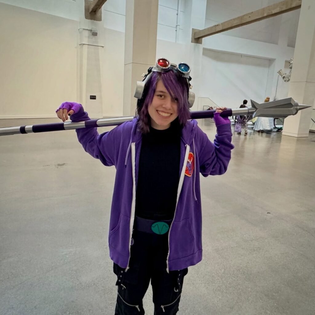A person in a purple costume holding a prop spear at an indoor event, wearing goggles on their head.