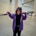 A person in a purple costume holding a prop spear at an indoor event, wearing goggles on their head.