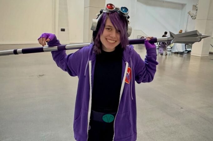 A person in a purple costume holding a prop spear at an indoor event, wearing goggles on their head.
