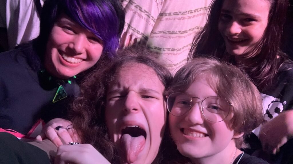 A group of young fans makes playful faces at a concert, with one sticking out their tongue.