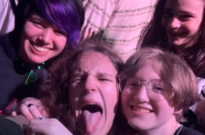 A group of young fans makes playful faces at a concert, with one sticking out their tongue.