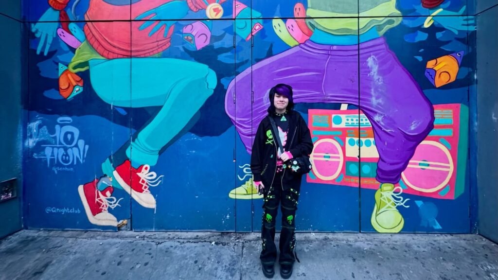A person stands before a colorful mural featuring dancing figures and a boombox. The artwork is bold with bright purple, blue, and red hues.
