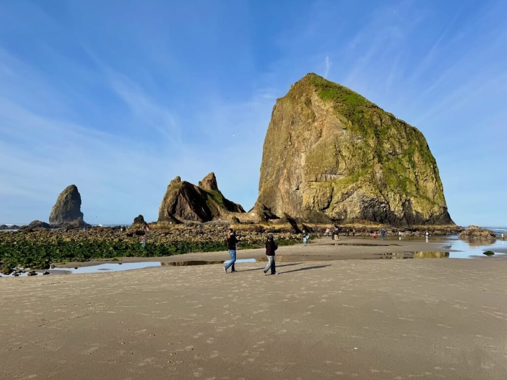 Visitors walking on the wide sandy beach near the massive Haystack Rock sea stack under a clear blue sky.