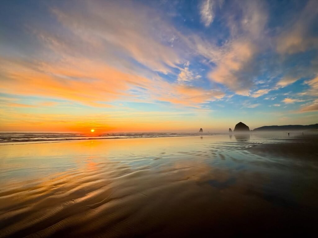 A serene sunset at a Pacific Northwest beach with warm orange and blue skies reflecting on wet sand. Scattered clouds add texture above the calm shoreline.