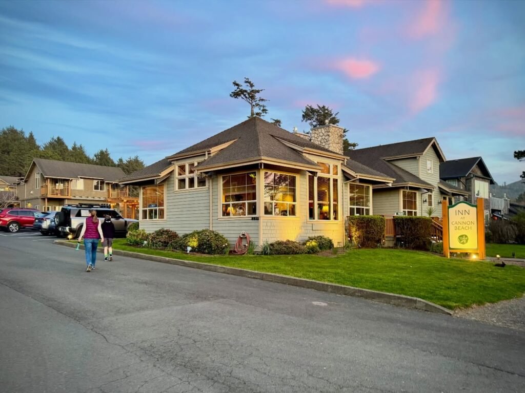 A cozy two-story home in the Pacific Northwest captures a stunning sunset sky with soft pink clouds. Pedestrians stroll on the street in front of the property.