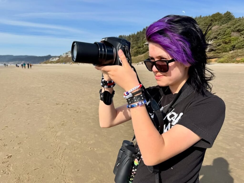 A person with purple hair holds a camera on a sunny beach. The background features sand, dunes, and a clear blue sky.
