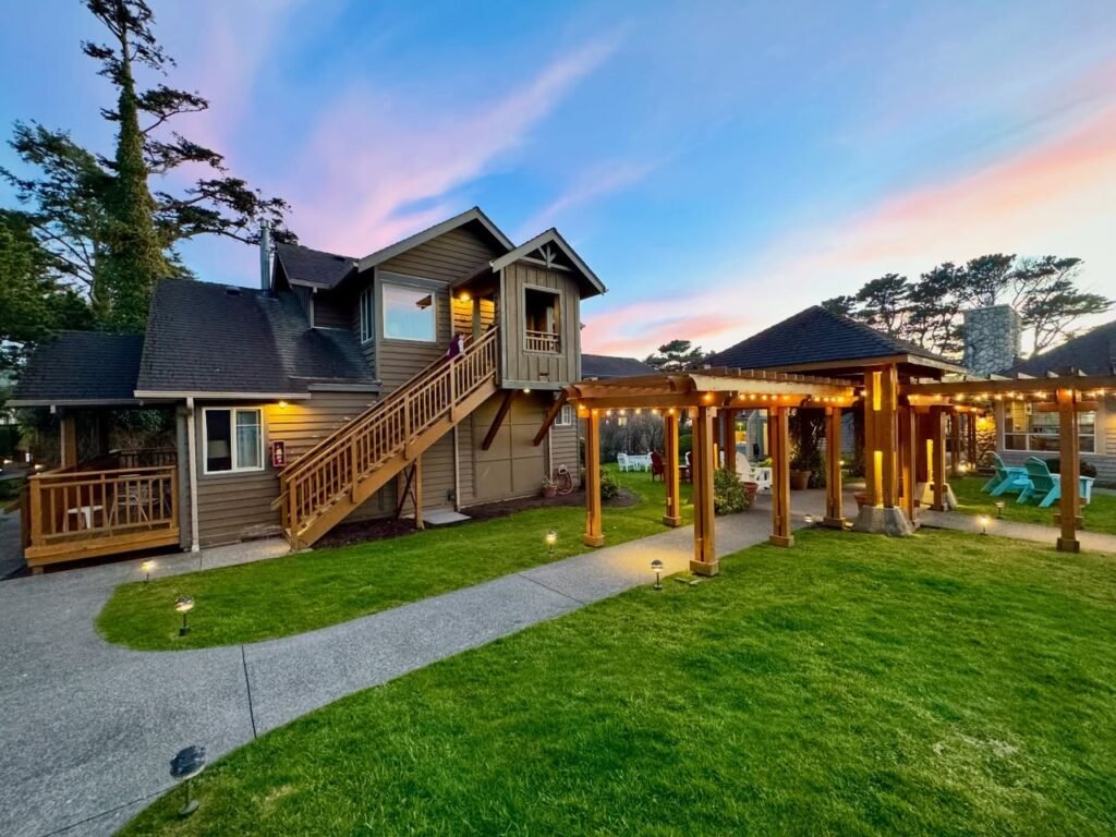 A two-story wooden lodge featuring a lit pergola and manicured lawns under a colorful twilight sky in the Pacific Northwest.