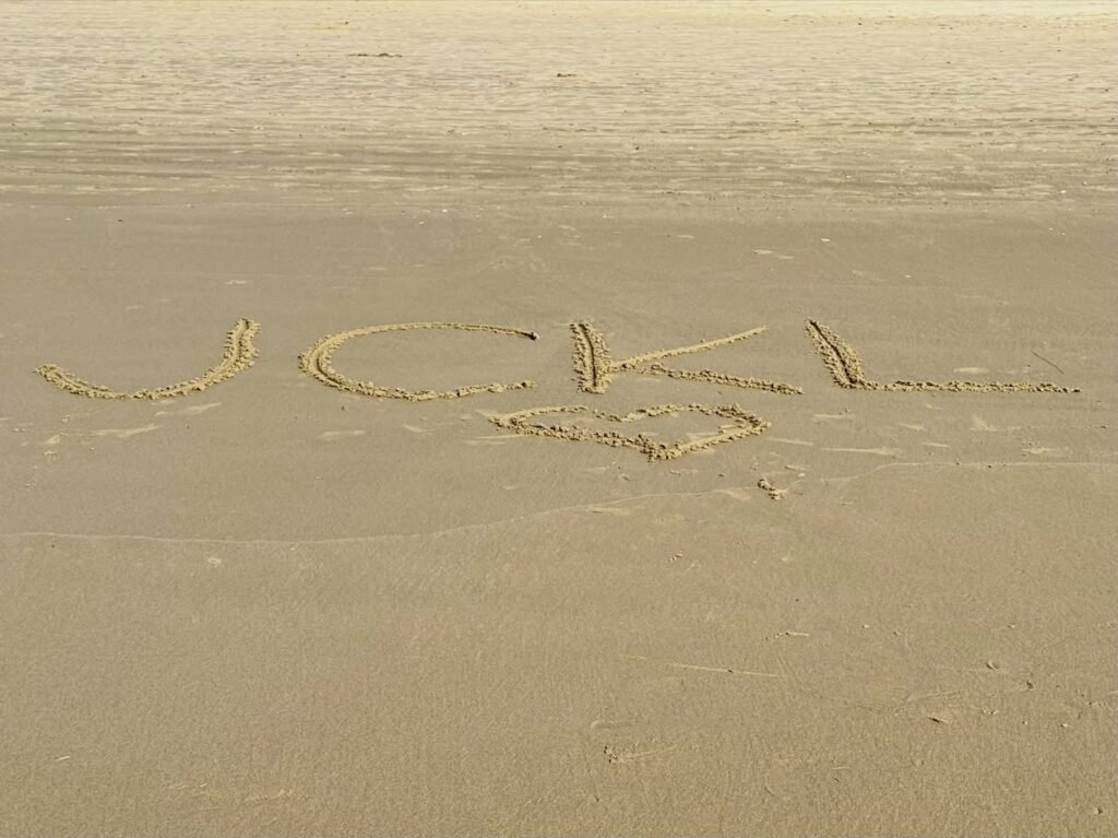 Letters spelling a name in wet sand on a beach near calm water.