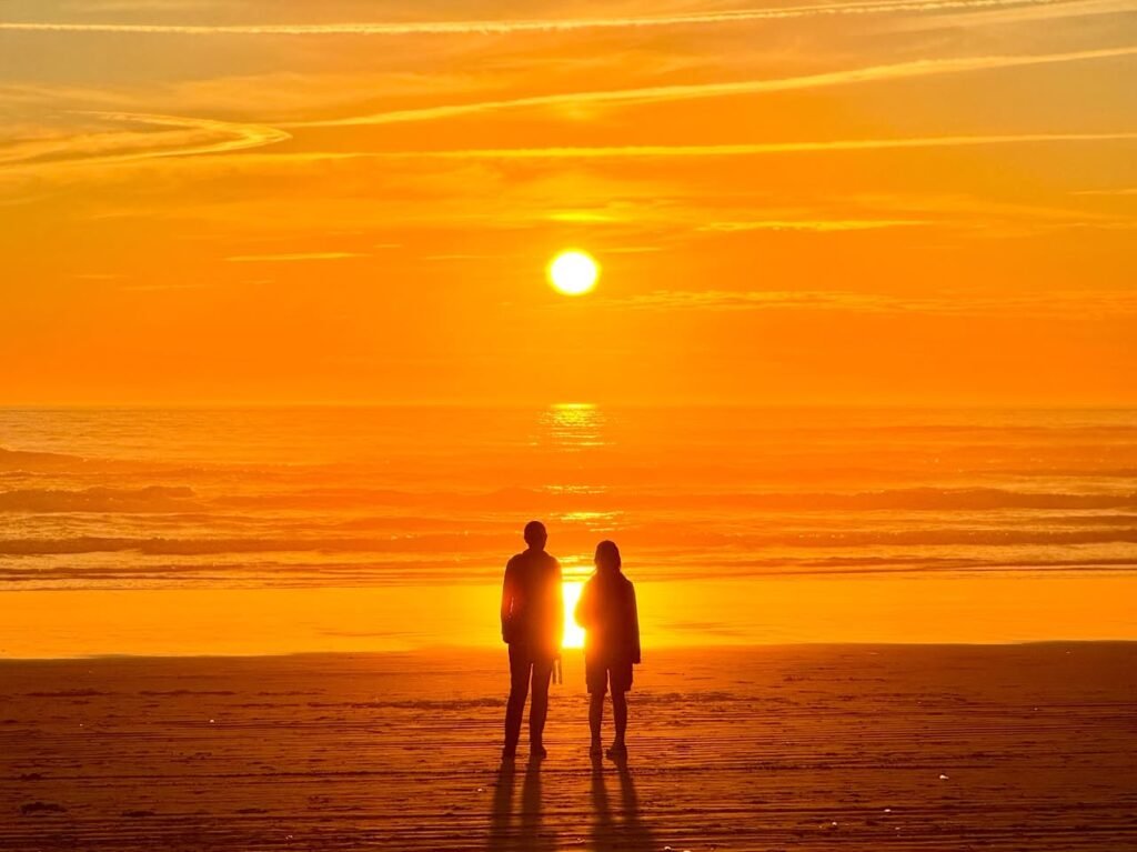 Two people stand silhouetted on a beach, facing a vibrant orange sunset over the ocean. The sun gleams behind them, casting a warm glow across the water and sky.