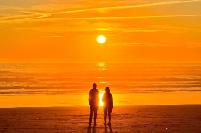 Two people stand silhouetted on a beach, facing a vibrant orange sunset over the ocean. The sun gleams behind them, casting a warm glow across the water and sky.