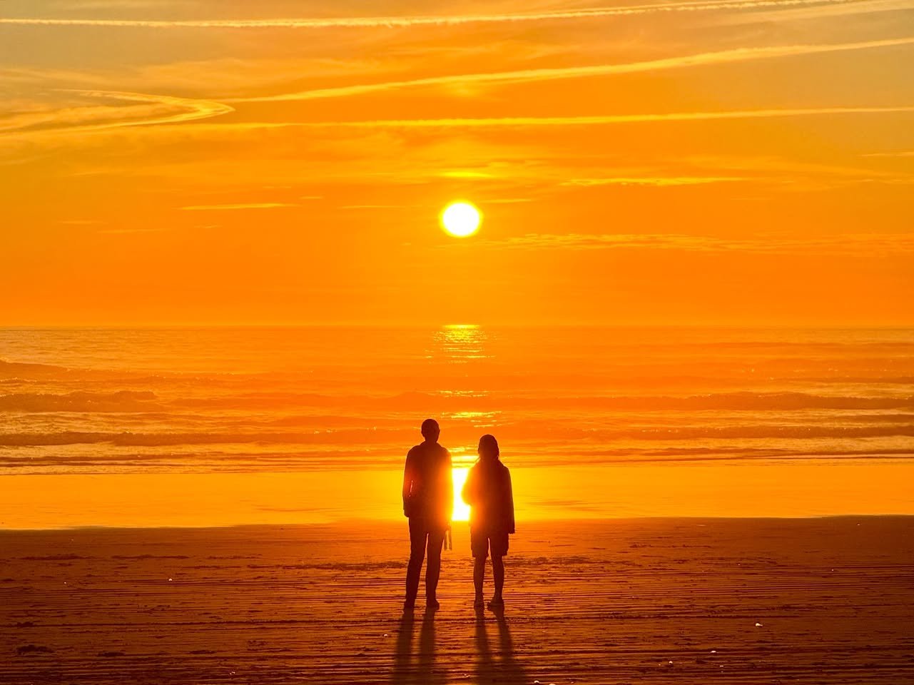 Two people stand silhouetted on a beach, facing a vibrant orange sunset over the ocean. The sun gleams behind them, casting a warm glow across the water and sky.