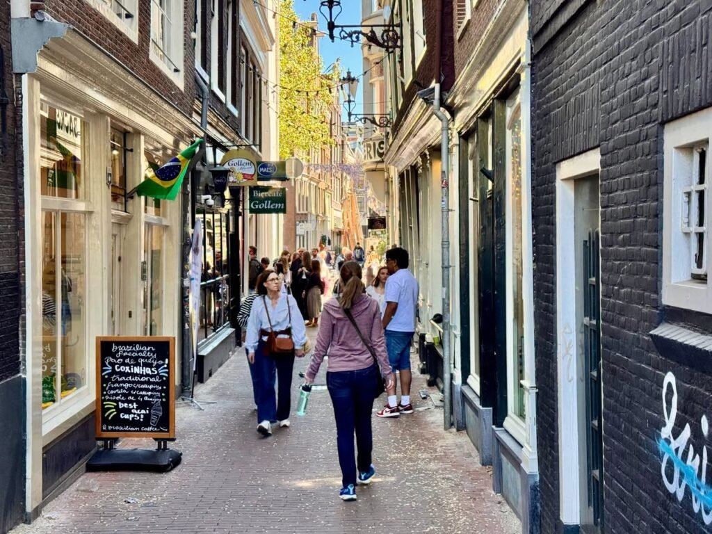 Travelers stroll through a narrow cobbled street in Edinburgh, with shop fronts, a chalkboard sign, and a Brazilian flag.