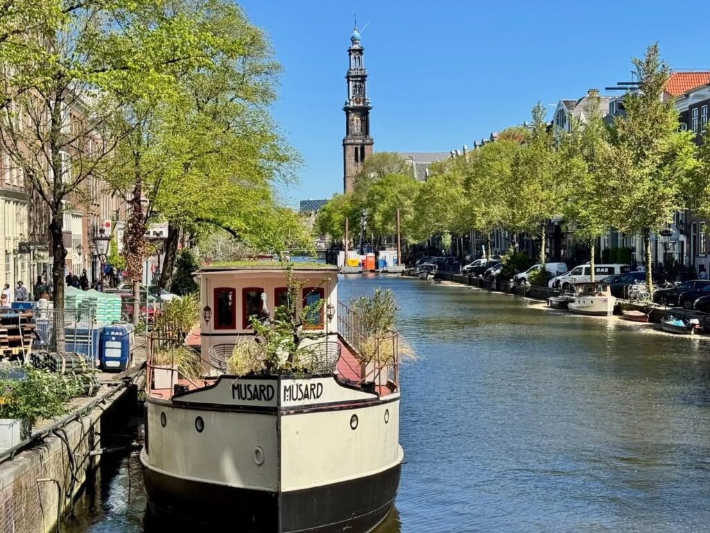 A canal barge named HUSARD passes through Bruges under a tall stone tower with lush green trees lining the waterways.