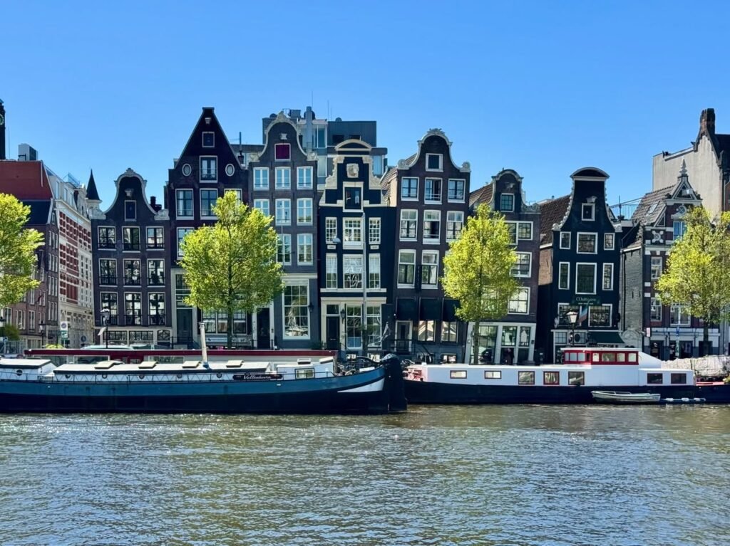 Traditional Dutch canal houses line the waterway with historic boats docked nearby under a clear blue sky. The scene captures the vibrant waterfront architecture of Amsterdam.
