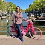 Person posing with a bicycle decorated in bright pink flowers beside a canal.