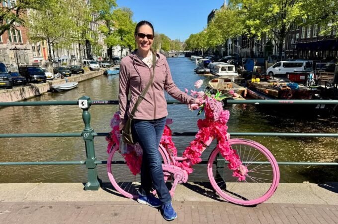 Person posing with a bicycle decorated in bright pink flowers beside a canal.