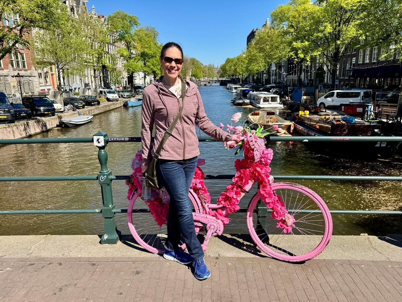 Person posing with a bicycle decorated in bright pink flowers beside a canal.