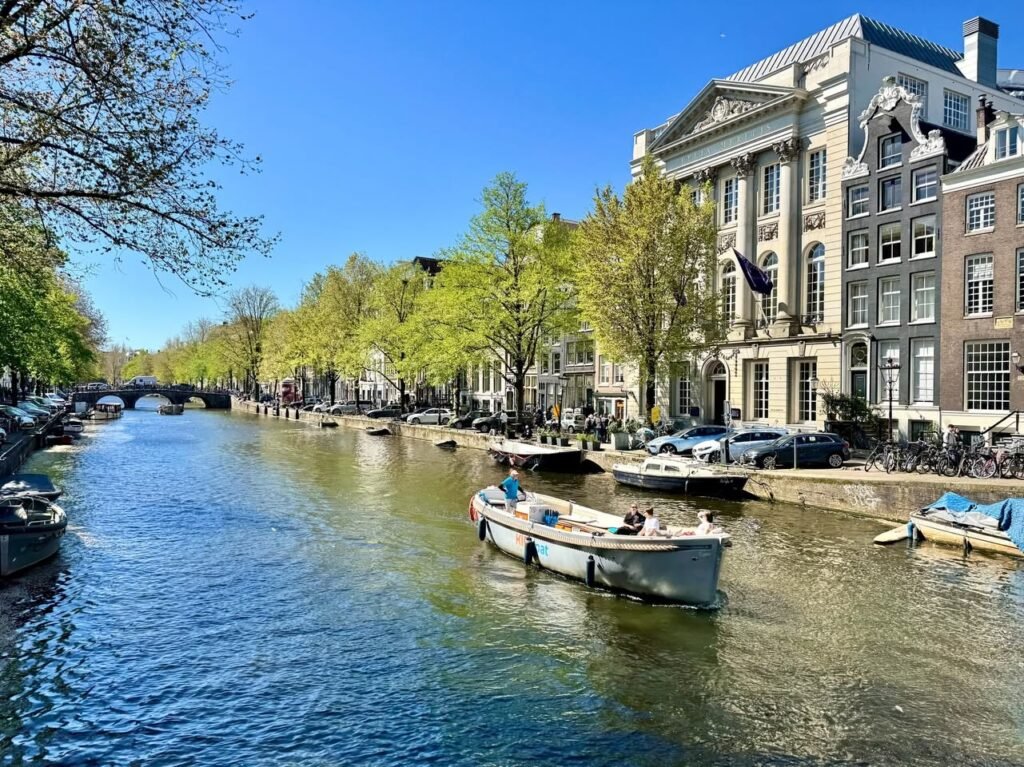 Sunny day on an Amsterdam canal with moored boats and historic buildings. Lush green trees line the waterway.