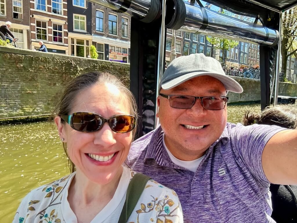A couple takes a selfie during a canal boat tour in Amsterdam, with historic buildings in the background.