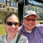 A couple takes a selfie during a canal boat tour in Amsterdam, with historic buildings in the background.