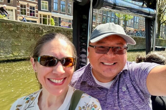 A couple takes a selfie during a canal boat tour in Amsterdam, with historic buildings in the background.