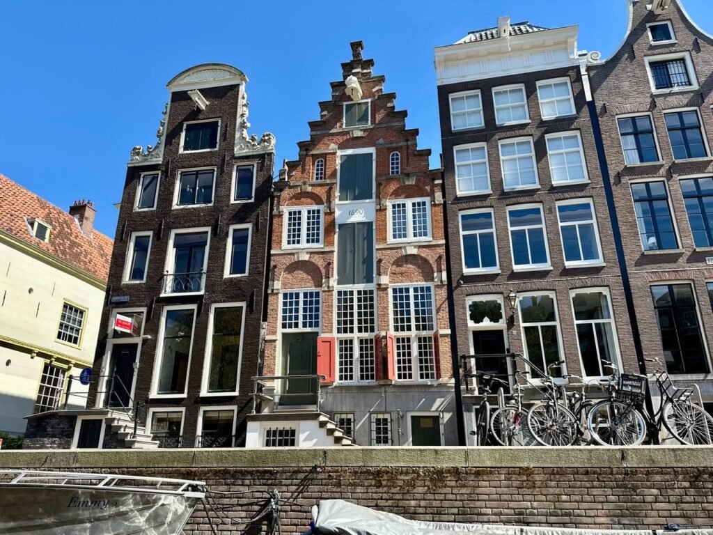 A row of traditional Dutch canal houses with unique gabled roofs, bicycles parked along the canal. Bright blue sky in the background.