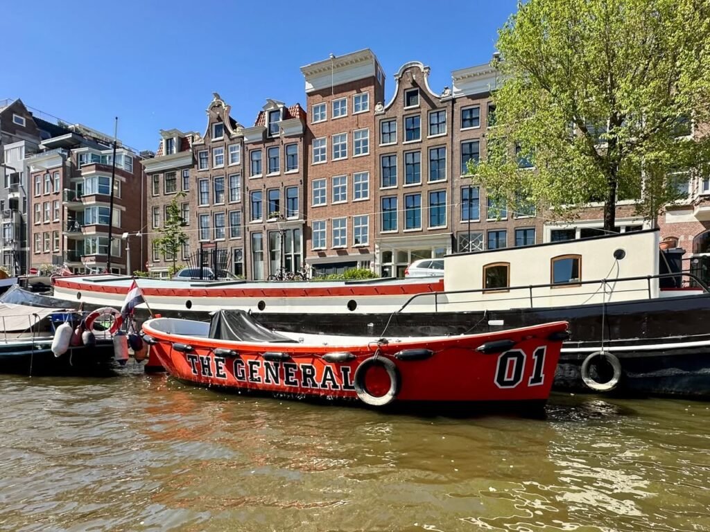 A red fireboat named 'THE GENERAL' floats on an Amsterdam canal, with historic gabled buildings lining the waterway under a clear blue sky.